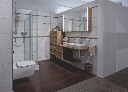 Ausstellungsbad mit großzügiger Glasdusche, wandhängendem WC, modernem Waschtisch aus Holz und großem Spiegelschrank.