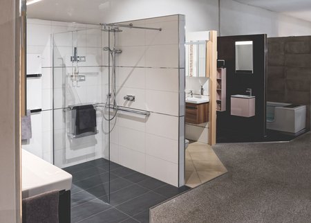 Badeausstellung mit bodengleicher Dusche, großem Glasabtrennungssystem, hellen Wandfliesen und zwei weiteren Waschtischbereichen im Hintergrund.