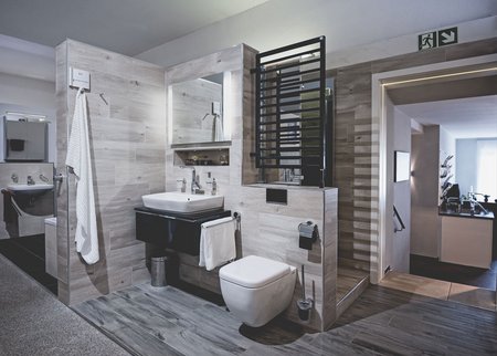 Badausstellung mit hellen Holzfliesen, wandhängendem Waschtisch, beleuchtetem Spiegel, schwarzem Design-Heizkörper und wandhängendem WC.