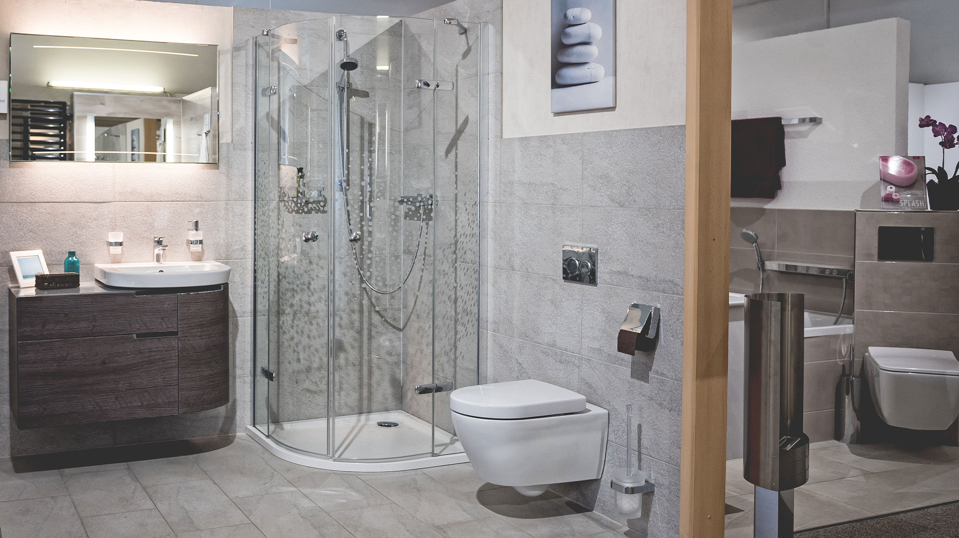 Modern eingerichtete Badausstellung mit Glas-Eckdusche, wandhängendem WC, dunklem Waschtischmöbel, beleuchtetem Spiegel und grauen Wandfliesen.